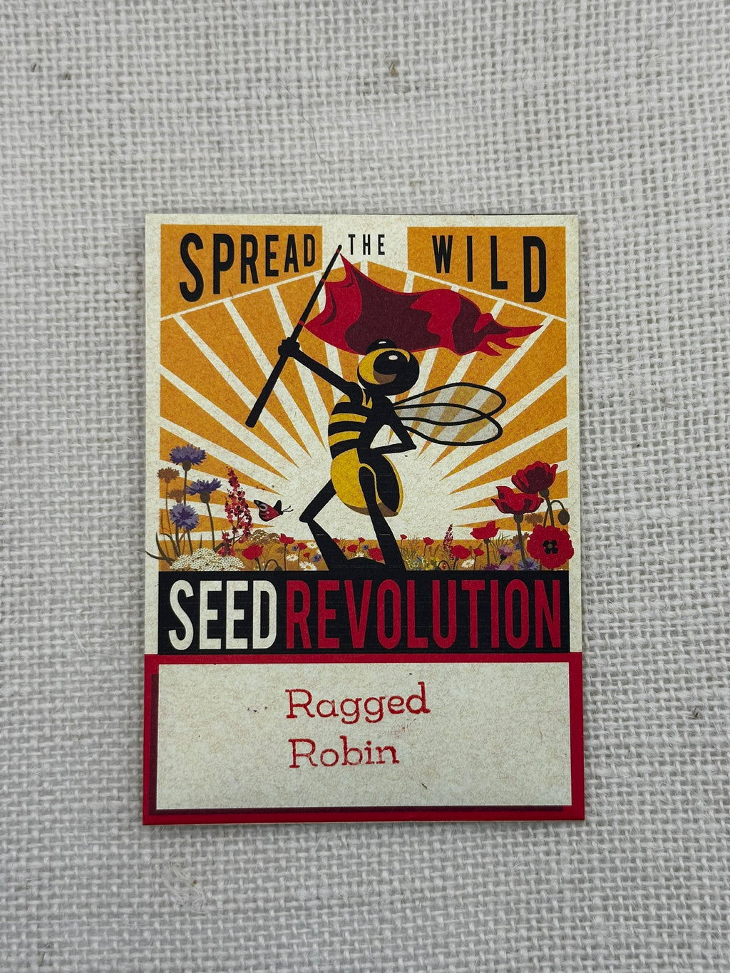 Seed packet design with bee and flag, text 'Spread the Wild Seed Revolution Ragged Robin' on a textured gray background.