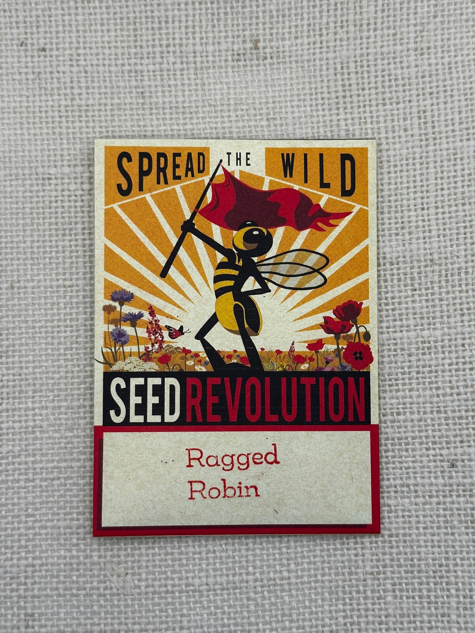 Seed packet design with bee and flag, text 'Spread the Wild Seed Revolution Ragged Robin' on a textured gray background.